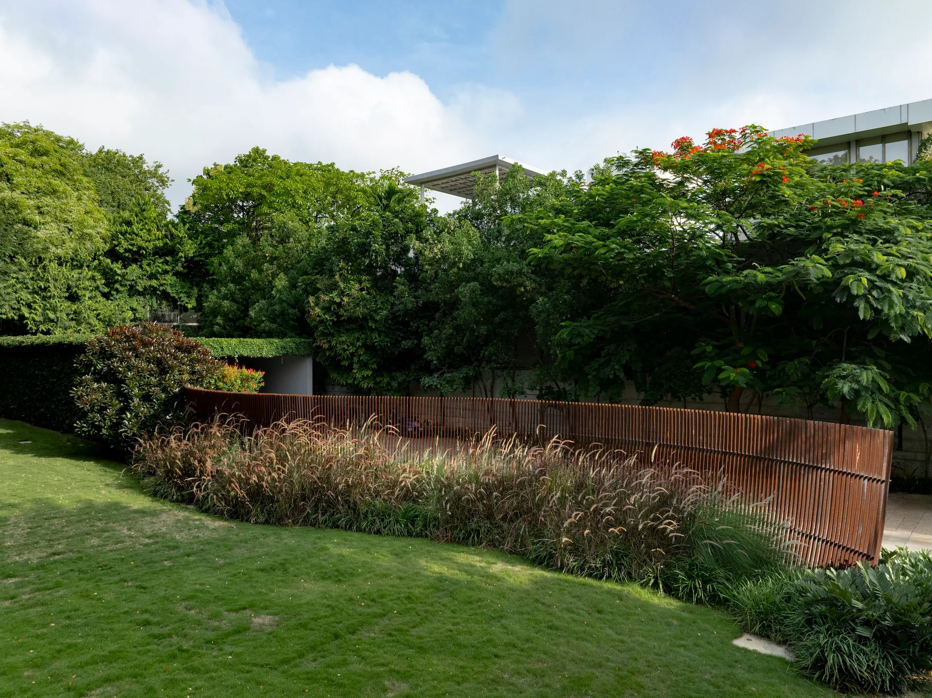 A curved corten steel wall runs along a lawn, with ornamental grasses at its foot and old trees behind.