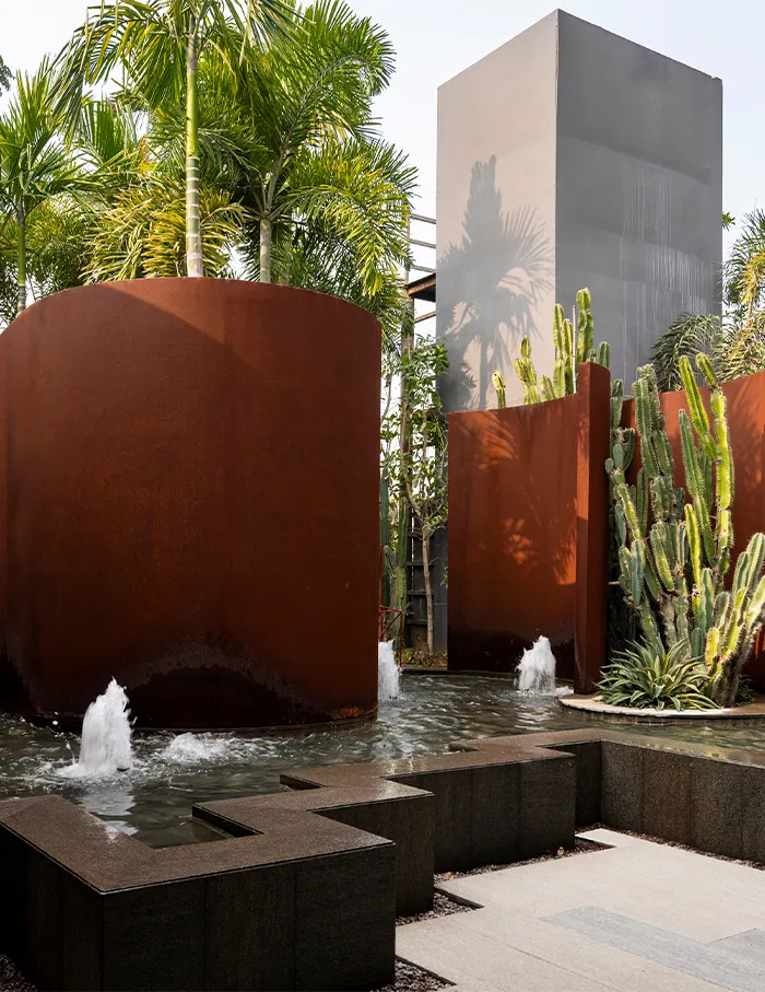 Tall cylinders of corten steel rise from a shallow pool with small fountains and a stand of cactus.