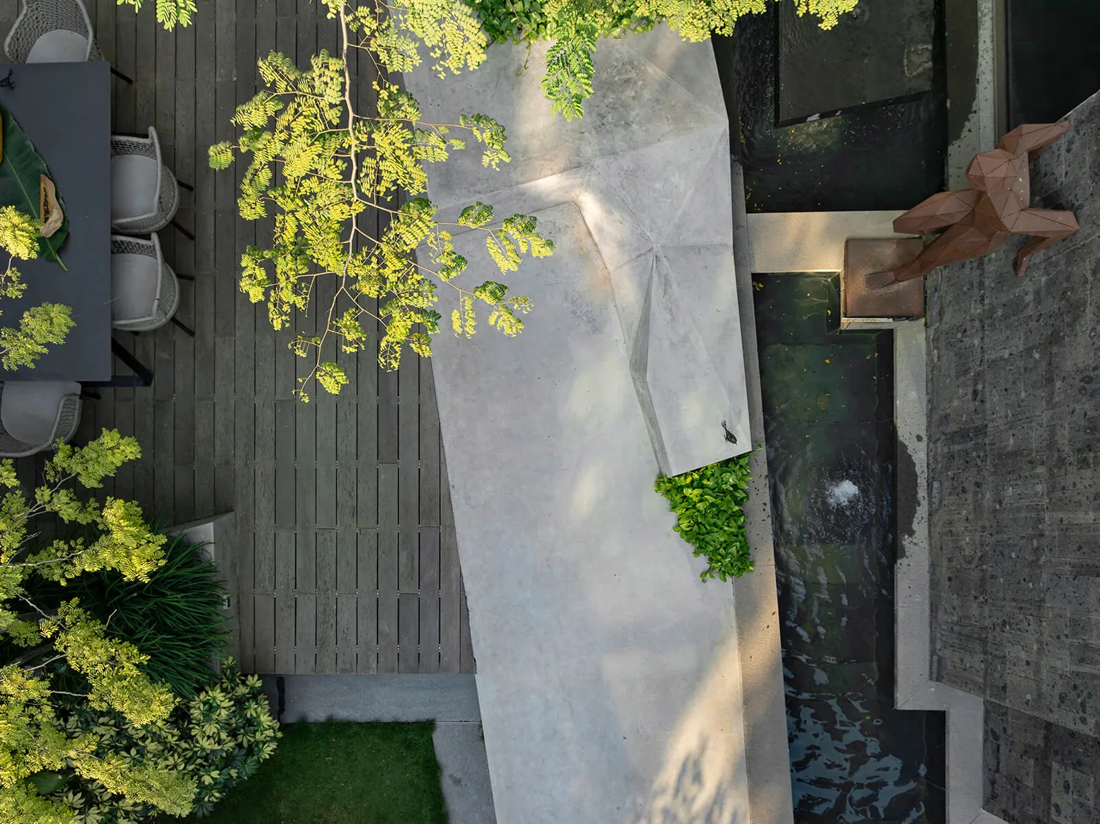 From above: a rust-coloured figure sculpture sits beside a narrow water channel and a pale stone path.
