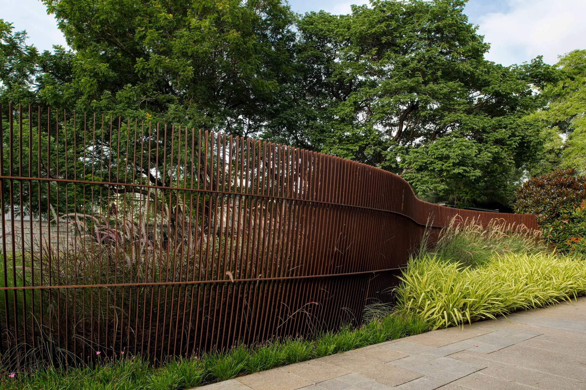 A slatted corten screen bends along a path, backed by grasses and trees.